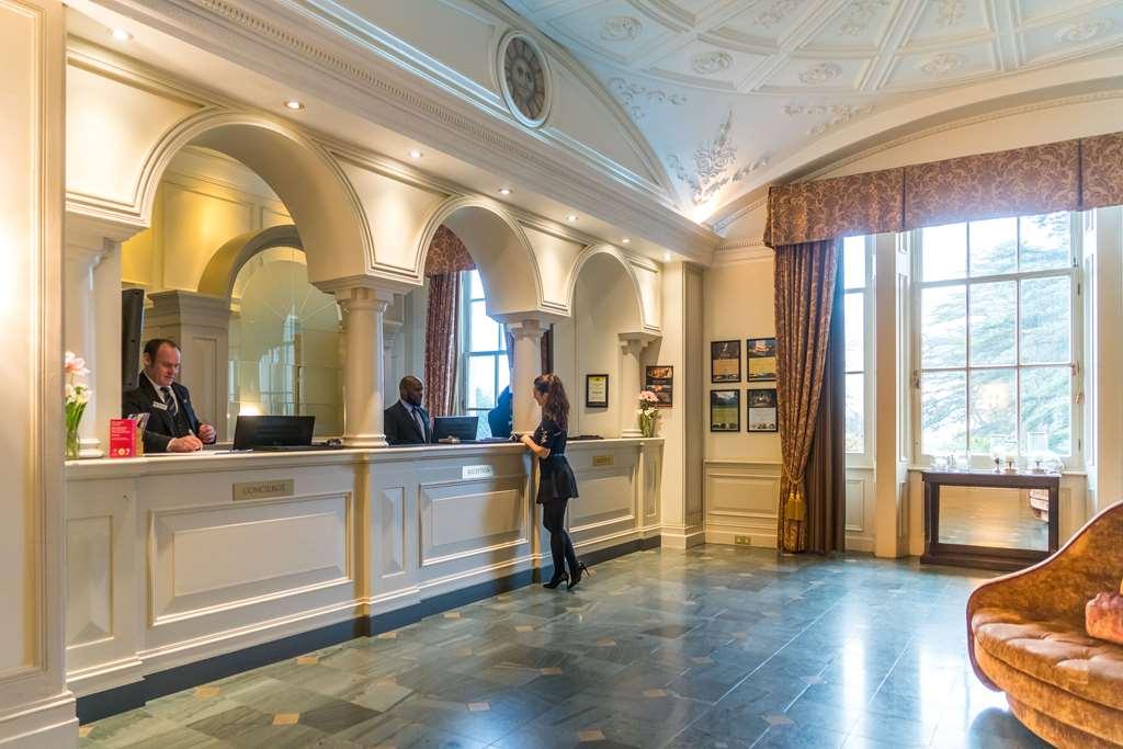 Macdonald Bath Spa Hotel Интериор снимка The photo shows the interior of a hotel reception area. There is a stylish front desk with several staff members attending to guests. A woman is interacting with one of the receptionists. The design features elegant architectural details, including a