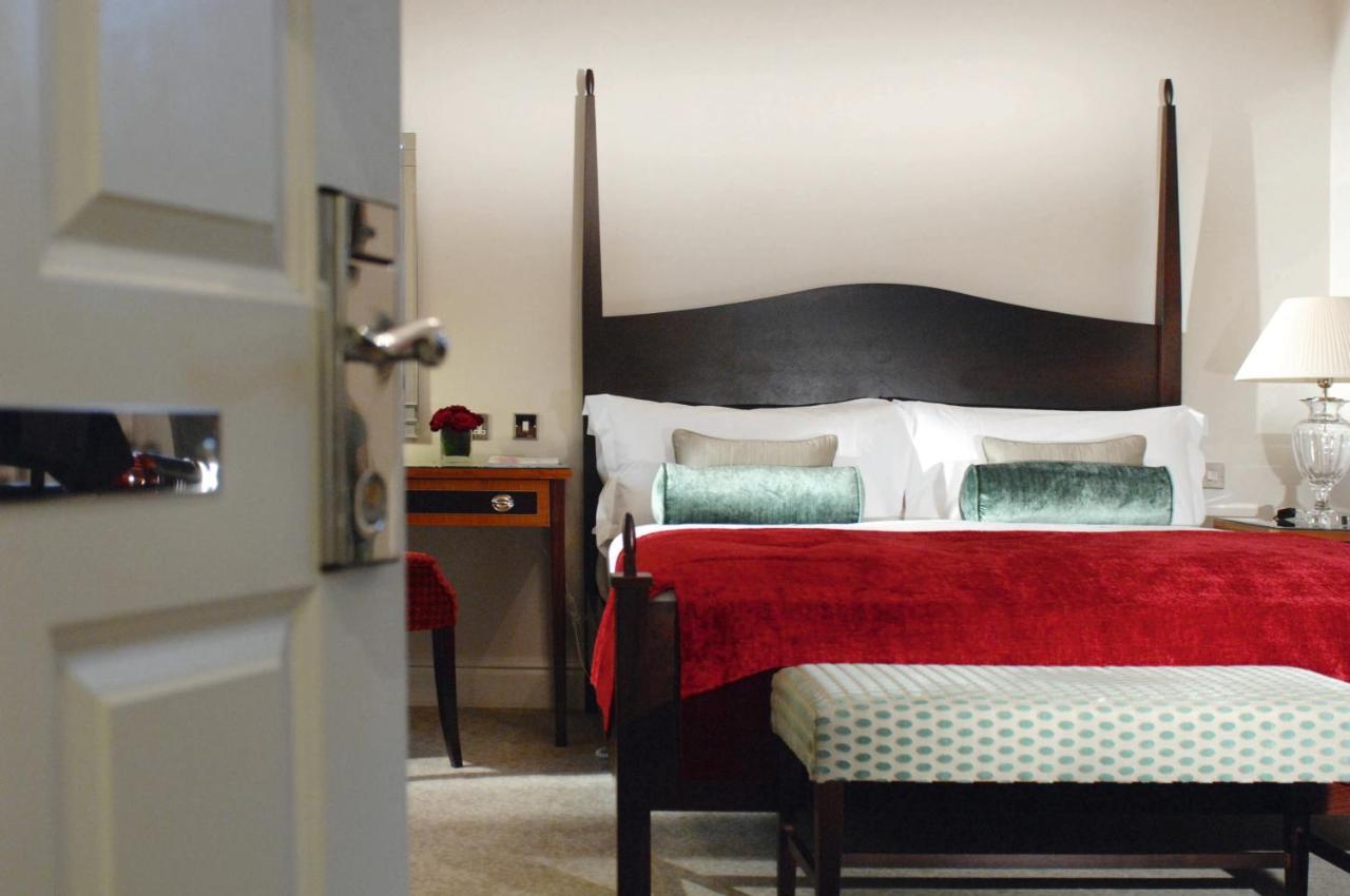Macdonald Bath Spa Hotel Стая снимка The photo shows a well-decorated hotel room. In the foreground, there is a partially opened door leading into the room. The focal point of the room is a large bed with a dark wooden headboard and stylish red and white bedding, accented with green pil