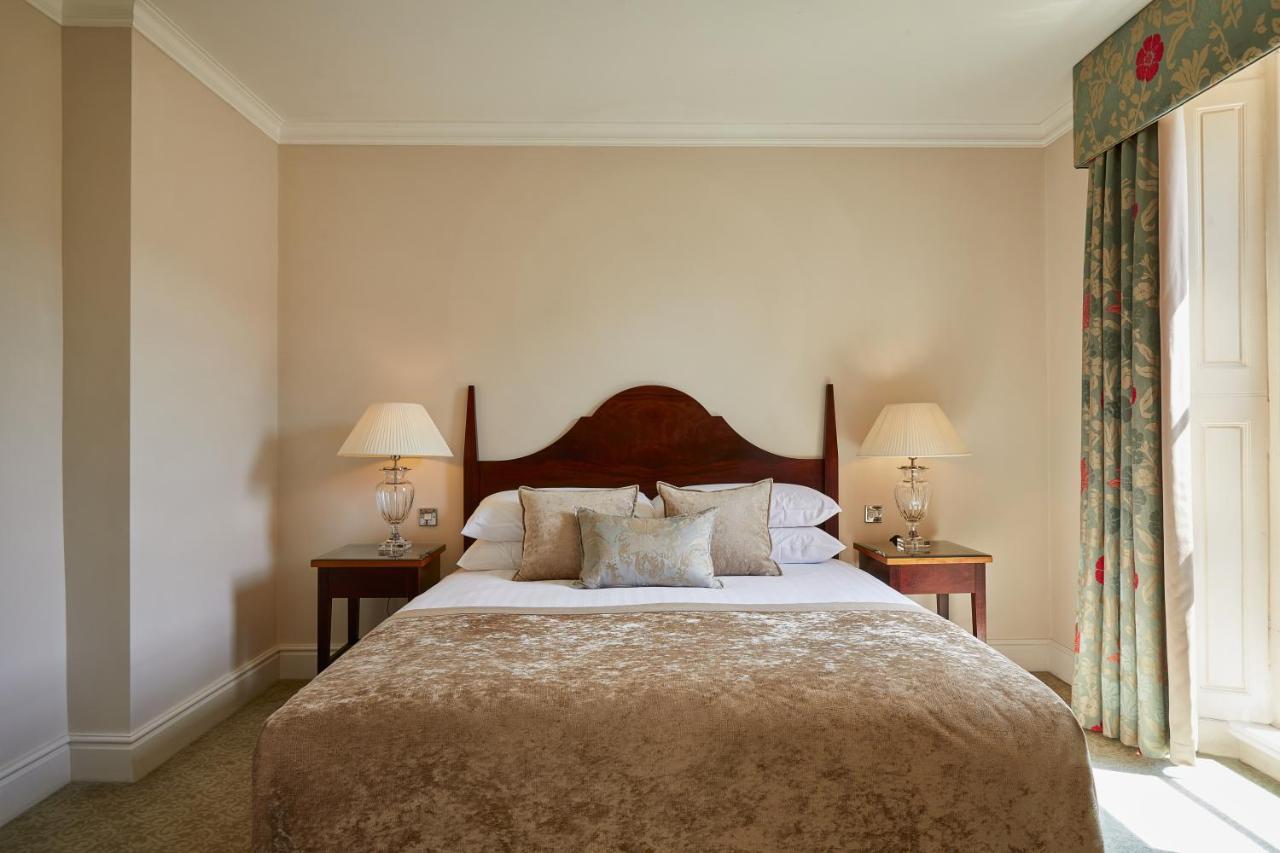Macdonald Bath Spa Hotel Екстериор снимка The photo shows a cozy hotel bedroom. It features a large bed with a plush, light-colored bedspread and several pillows. On either side of the bed, there are wooden nightstands with lamps providing soft lighting. The walls are painted a neutral color