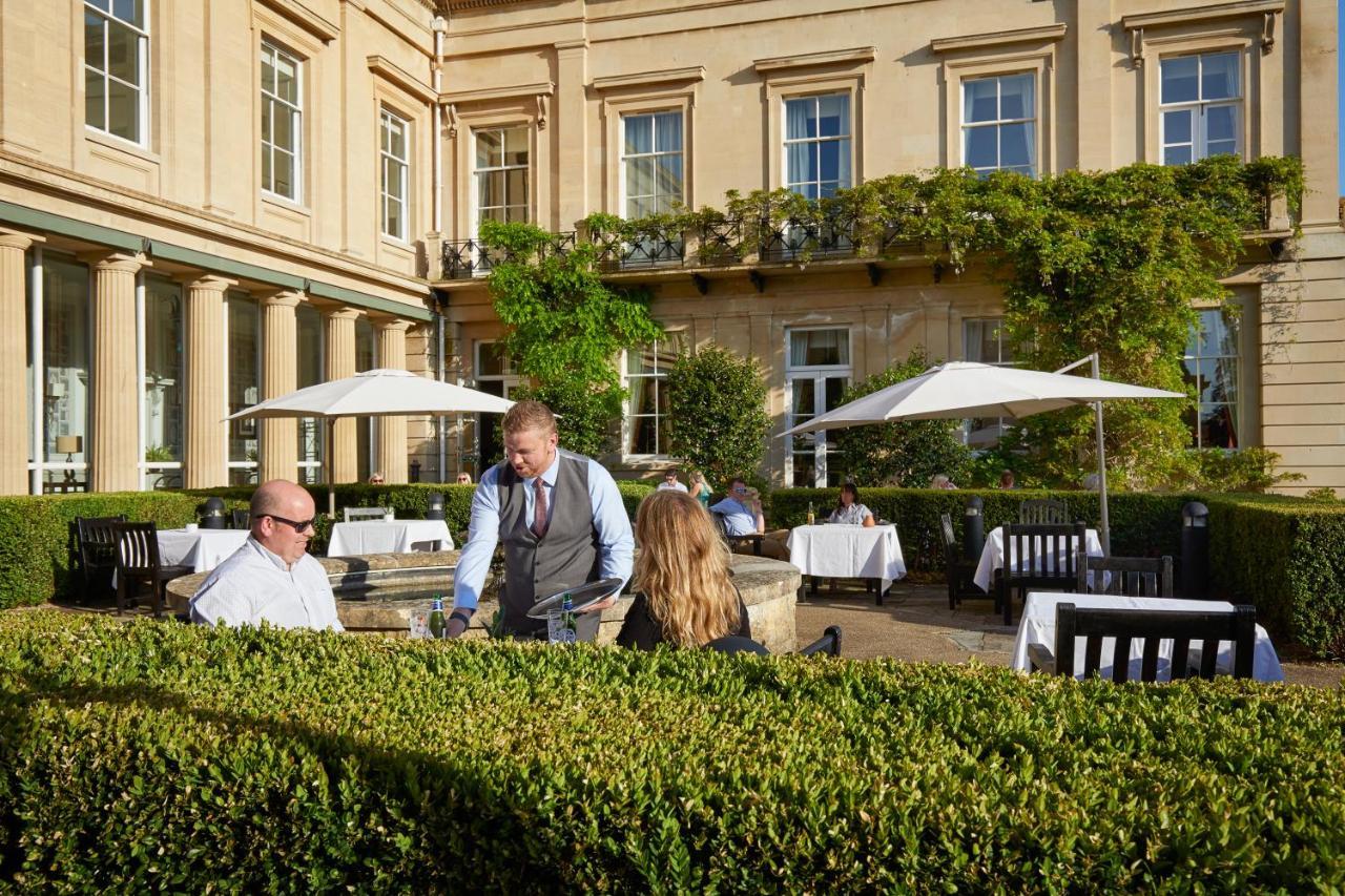 Macdonald Bath Spa Hotel Екстериор снимка The photo depicts an outdoor dining area of a restaurant. In the foreground, there are neatly trimmed hedges surrounding the dining space. Two patrons are seated at a table; one is a man wearing sunglasses and the other is a woman facing him. A waite