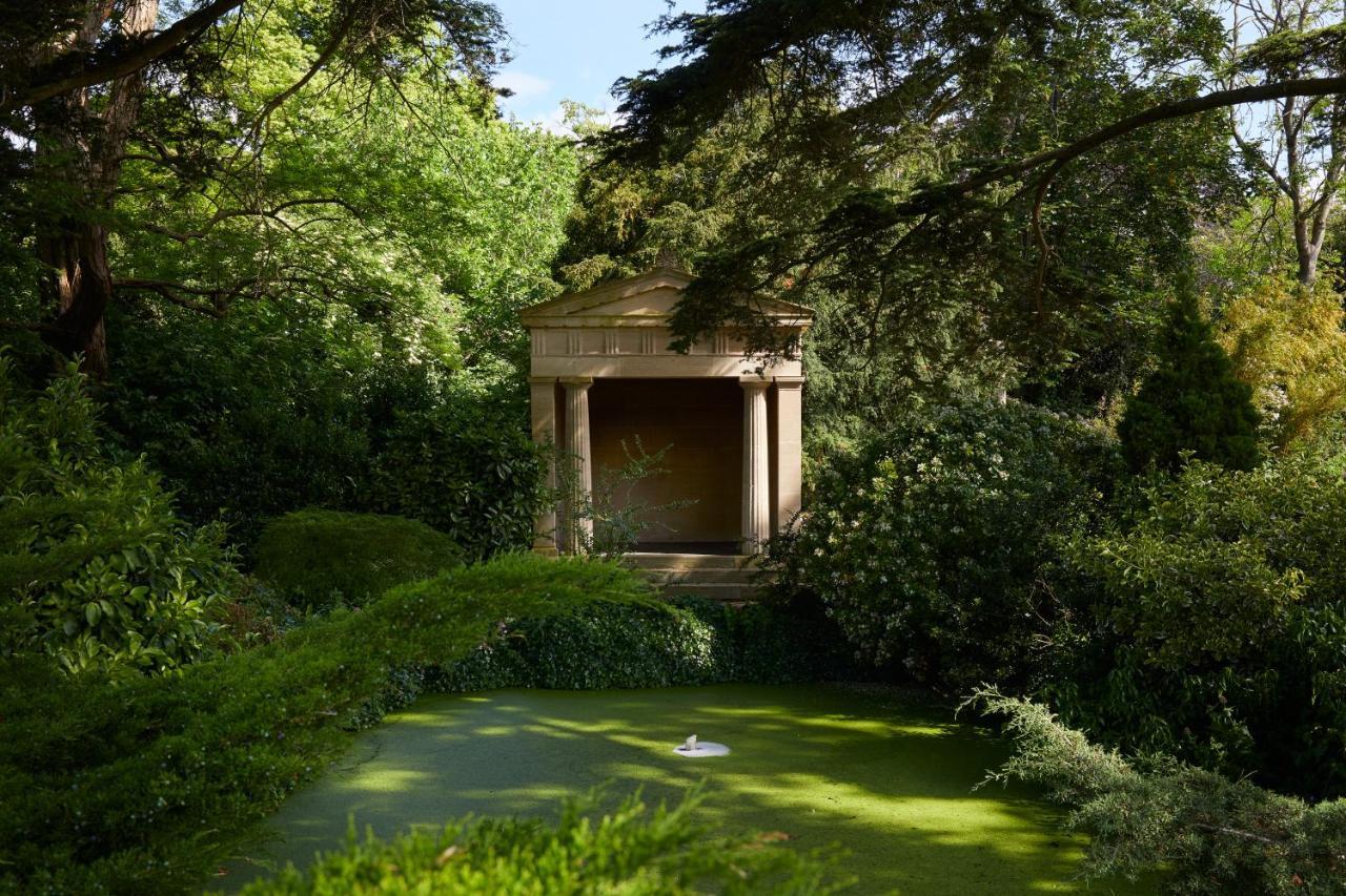 Macdonald Bath Spa Hotel Екстериор снимка The photo shows a serene garden scene featuring lush greenery and a tranquil pond. In the background, there is a classical-looking structure with columns, resembling a small temple or pavilion. The pond appears to have vegetation covering its surface