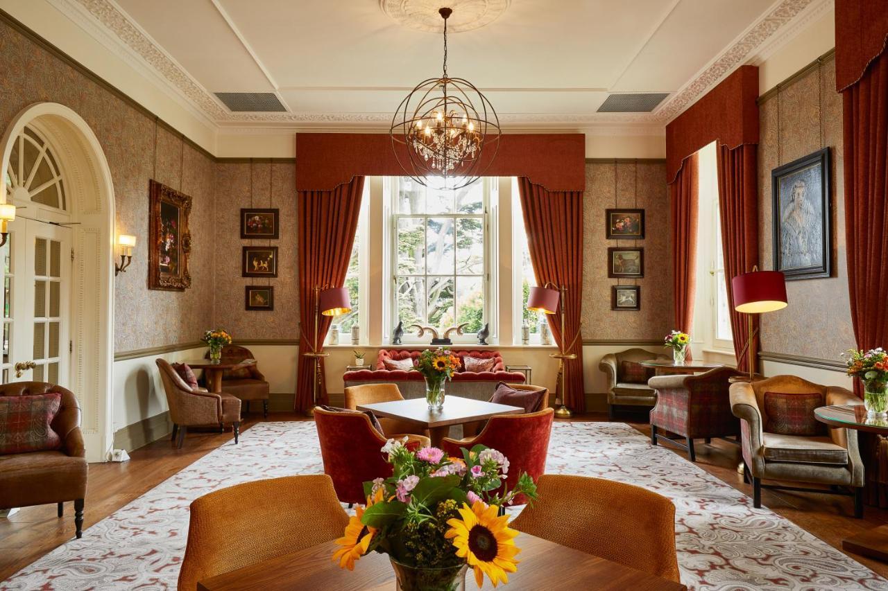 Macdonald Bath Spa Hotel Екстериор снимка The photo shows a well-decorated living room or lounge area. The space features elegant furnishings, including chairs and a round table at the center. Large windows allow natural light to fill the room, draped with rich red curtains. The walls are ad