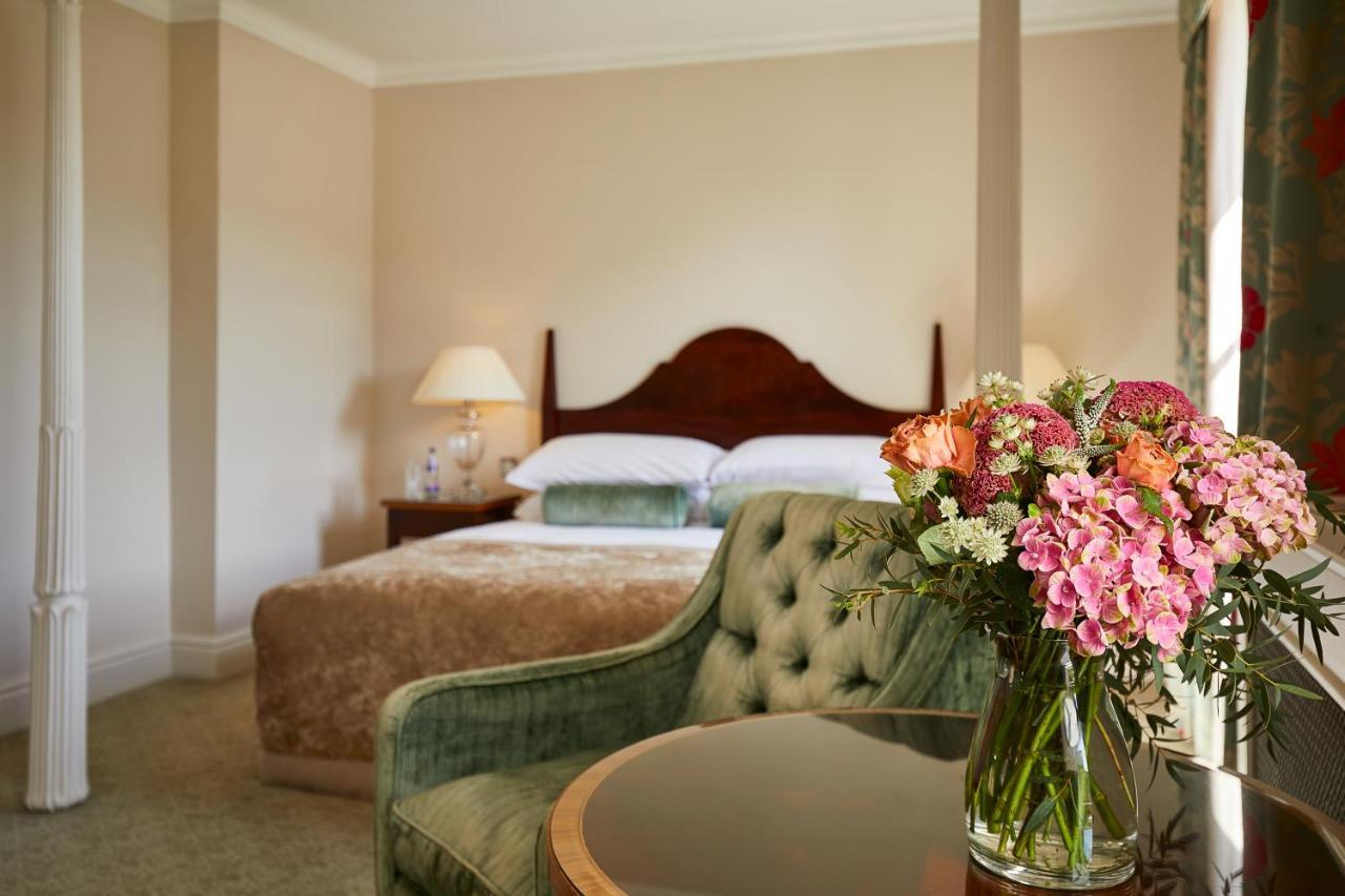 Macdonald Bath Spa Hotel Екстериор снимка The photo shows a cozy bedroom featuring a large bed with a wooden headboard, topped with a fluffy blanket. To one side, there is a small bedside lamp providing soft lighting. In the foreground, there is an elegant green chair with a tufted design an