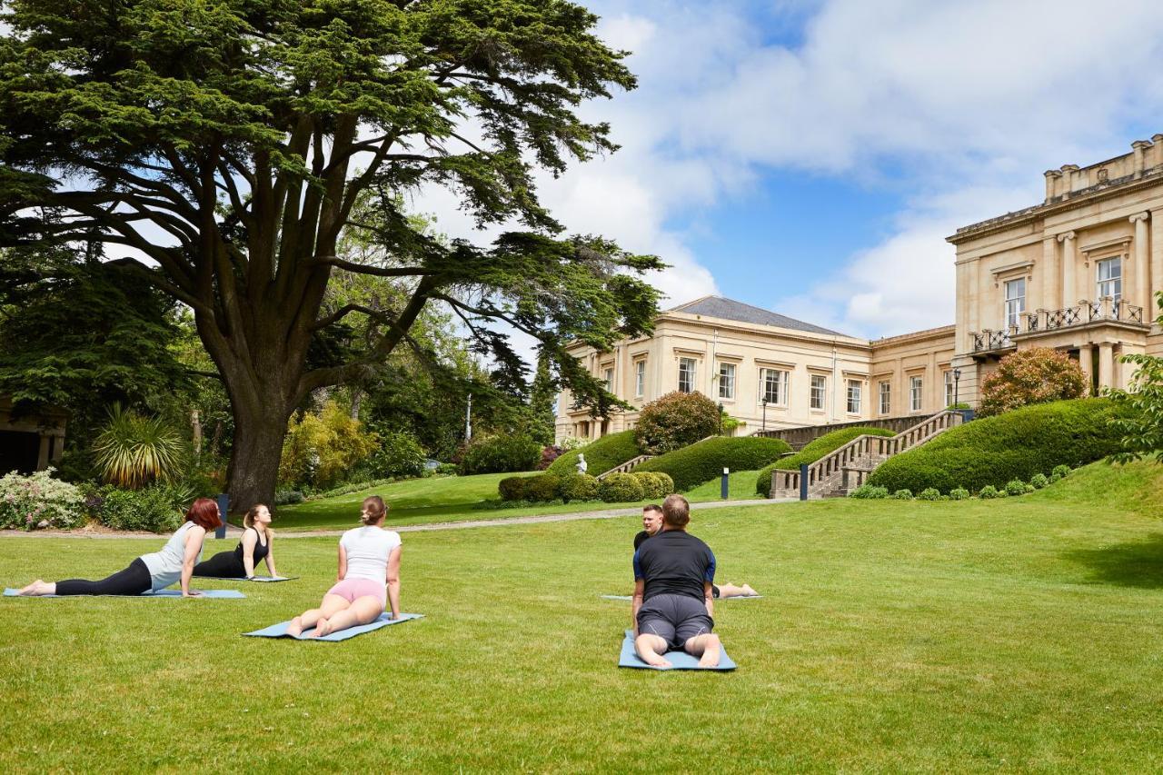 Macdonald Bath Spa Hotel Екстериор снимка The photo shows a group of people engaging in a yoga session outdoors. They are positioned on yoga mats on a lush green lawn in front of a large, elegant building with columns. Behind them, there are trees and well-maintained gardens, contributing to