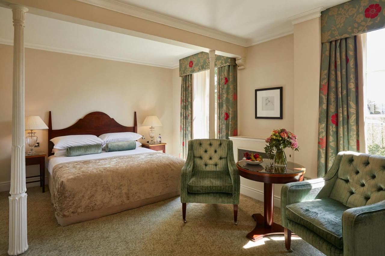 Macdonald Bath Spa Hotel Екстериор снимка The photo shows a cozy hotel room featuring a large bed with a beige bedspread. There are two green armchairs positioned next to a small round table. The decor includes floral curtains and a neutral color palette on the walls. Soft lighting is provid