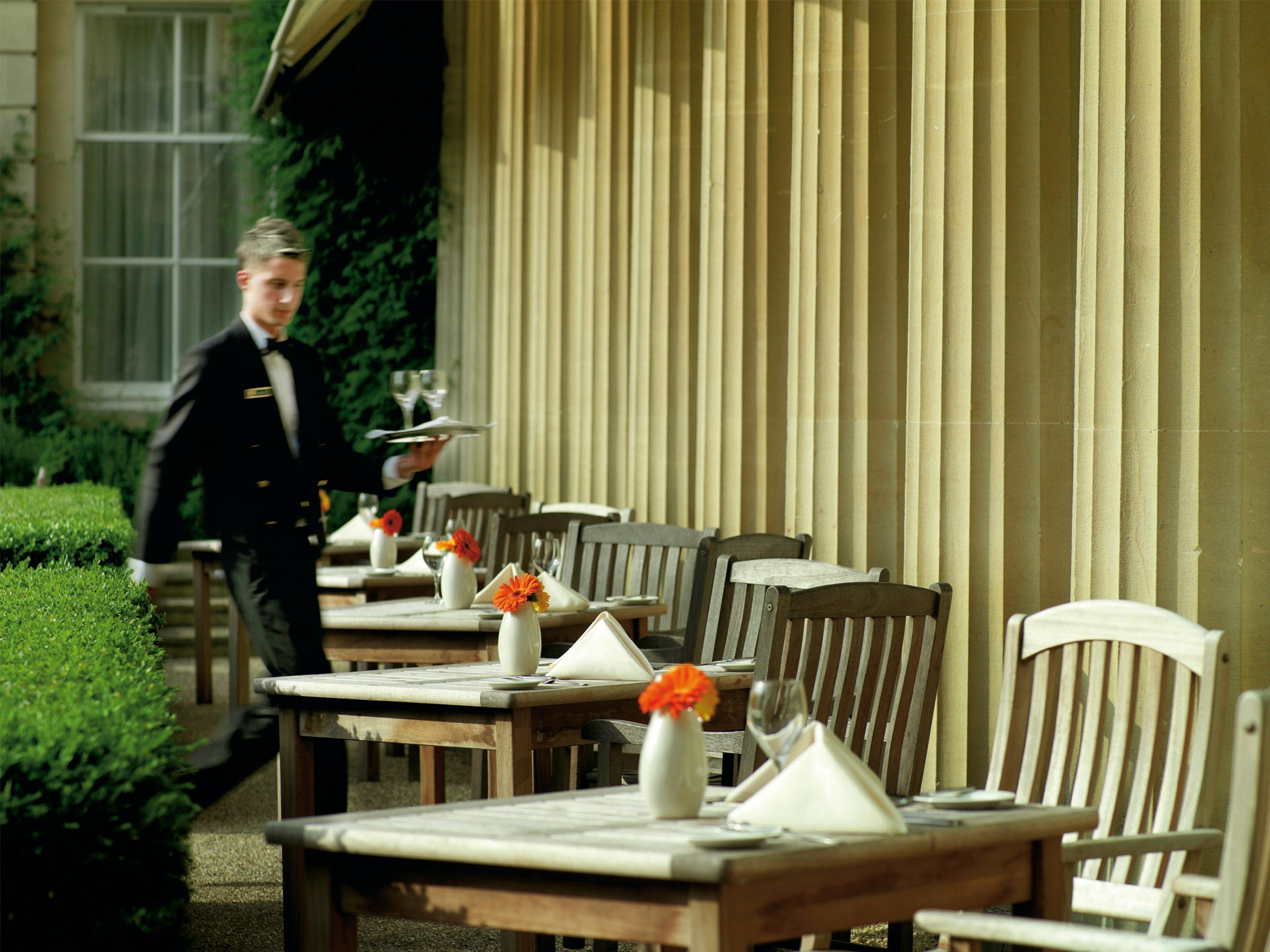 Macdonald Bath Spa Hotel Екстериор снимка The photo shows an outdoor dining area, featuring several wooden tables and chairs arranged neatly. Each table is set with a white tablecloth and small vases containing flowers, adding a vibrant touch. A waiter in a formal black suit is seen moving t