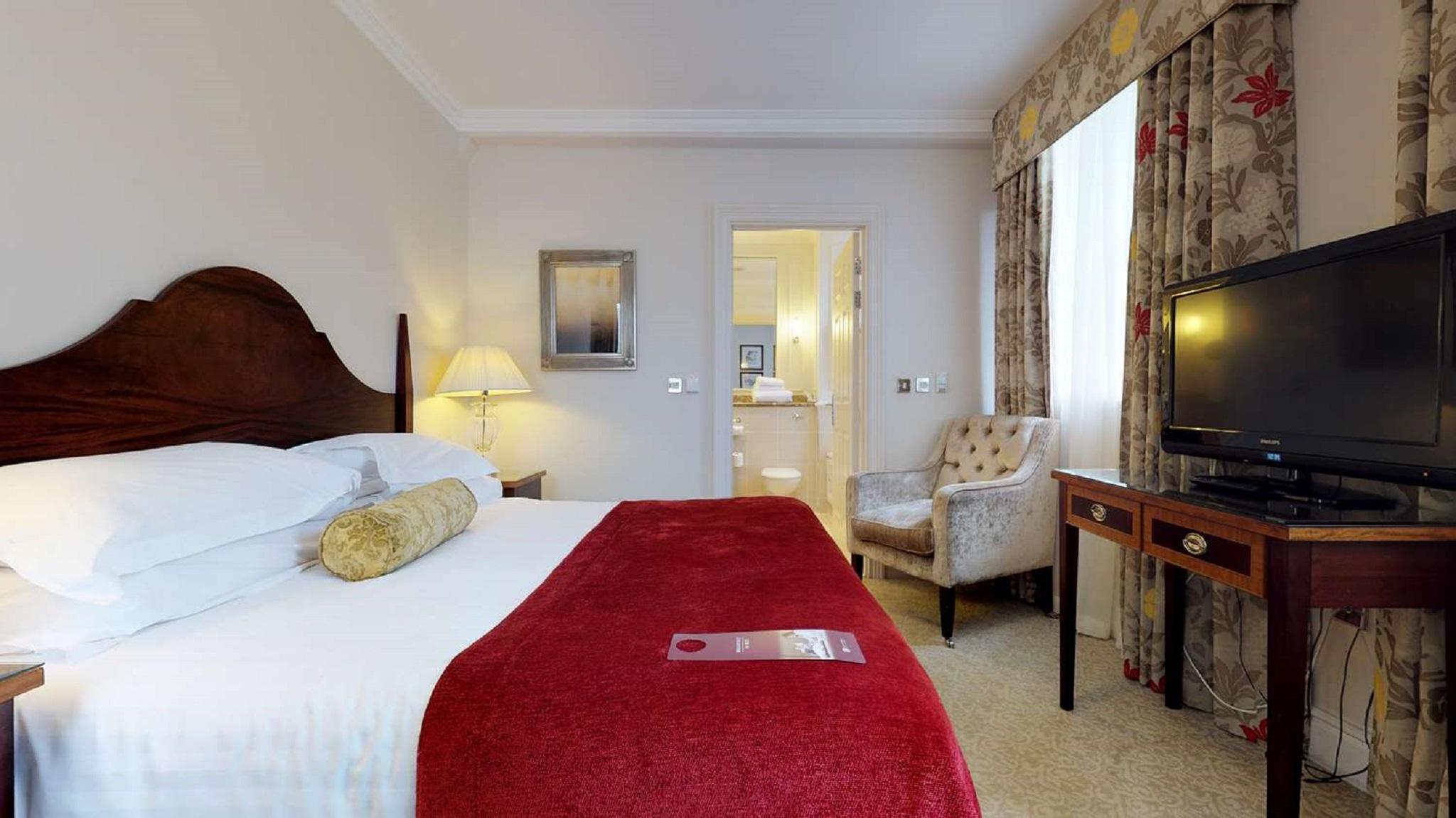 Macdonald Bath Spa Hotel Екстериор снимка The photo shows a hotel room with a queen-sized bed covered in white linens and a deep red throw blanket. There is a decorative pillow on the bed. Next to the bed, there's a small nightstand with a lamp. In the background, a seating area features a b