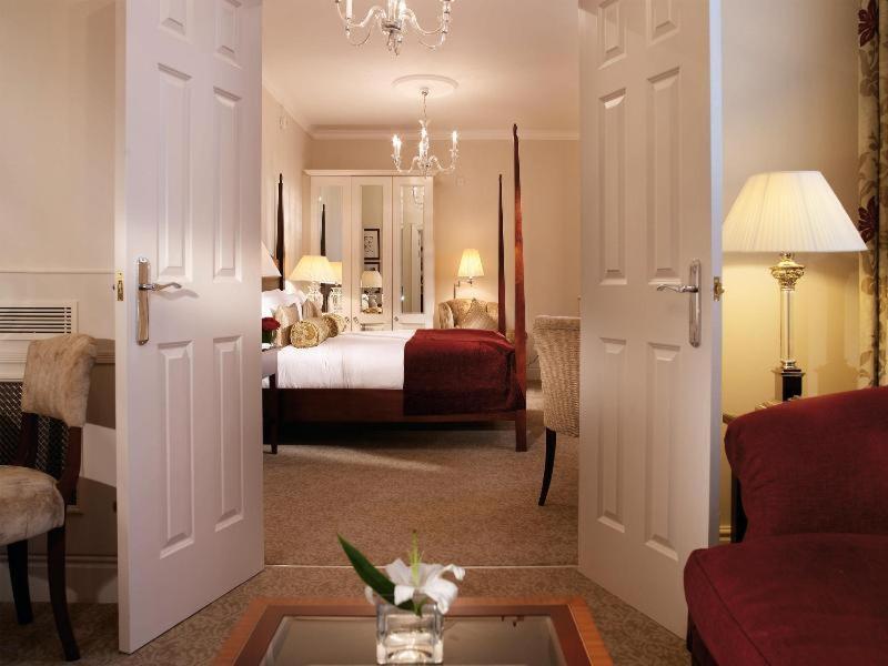 Macdonald Bath Spa Hotel Стая снимка The photo shows a hotel room that can be viewed through open double doors. Inside, there is a neatly made bed with decorative pillows, a plush area rug, and soft lighting from lamps. The room features elegant furnishings, including a chair, a coffee 