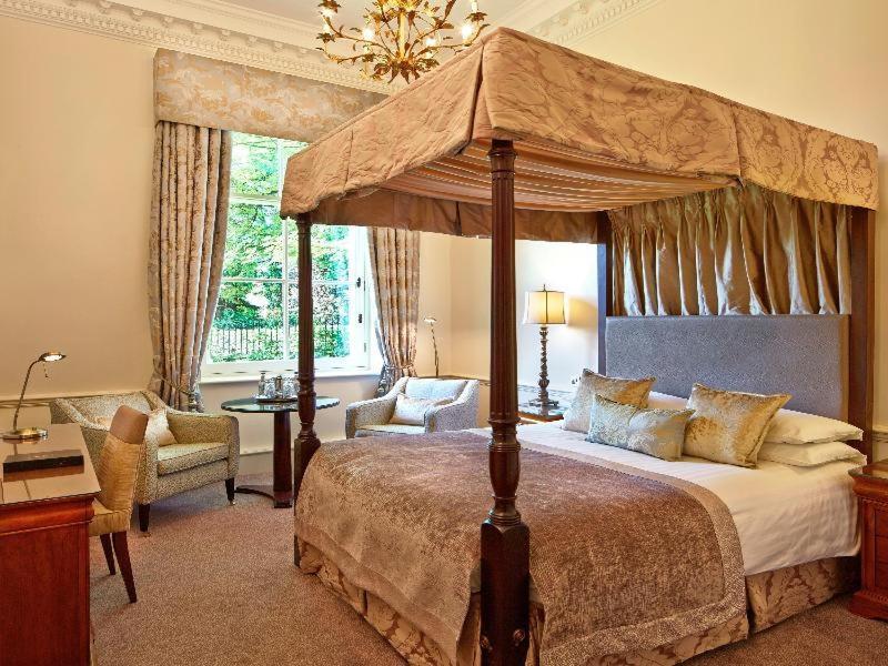 Macdonald Bath Spa Hotel Стая снимка The photo shows an elegantly designed bedroom featuring a four-poster bed with a decorative canopy. The bed is dressed with luxurious bedding and pillows, creating a cozy and inviting atmosphere. Flanking the bed are two comfortable armchairs and a s
