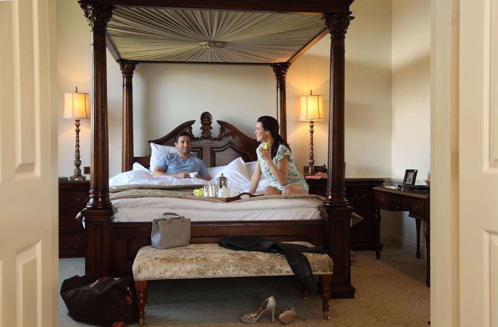 Macdonald Bath Spa Hotel Екстериор снимка The image depicts a spacious, well-lit bedroom featuring a large four-poster bed with an ornate headboard. A man and a woman are sitting on the bed, appearing to engage in conversation. The woman is dressed in a patterned outfit, while the man is wea