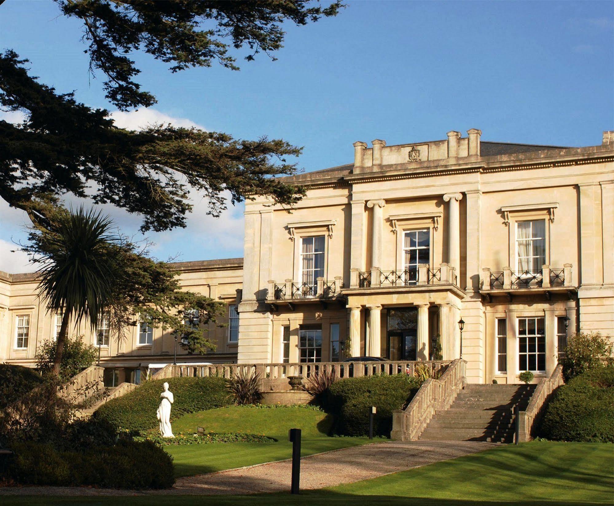 Macdonald Bath Spa Hotel Екстериор снимка The photo shows an elegant building with classical architecture, featuring large windows and decorative elements. The facade is light-colored, and there are several balconies. In front of the building, there is a landscaped area with greenery, includ