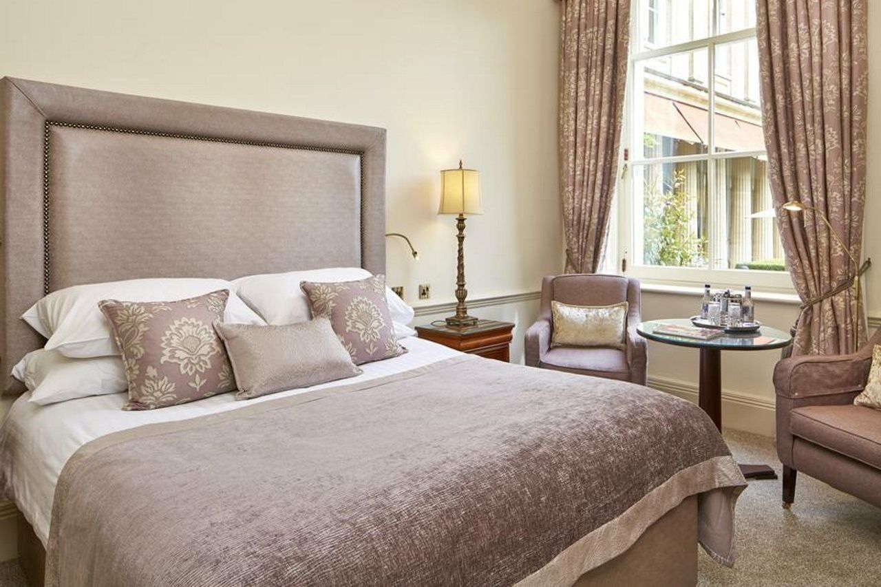 Macdonald Bath Spa Hotel Екстериор снимка The photo shows a stylish and elegantly decorated hotel room. It features a large bed with soft linens and several decorative pillows. The bedspread is a muted shade of beige or taupe. Beside the bed, there's a small table with two armchairs. One of 